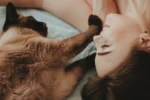 A Siamese cat lies next to a smiling woman.